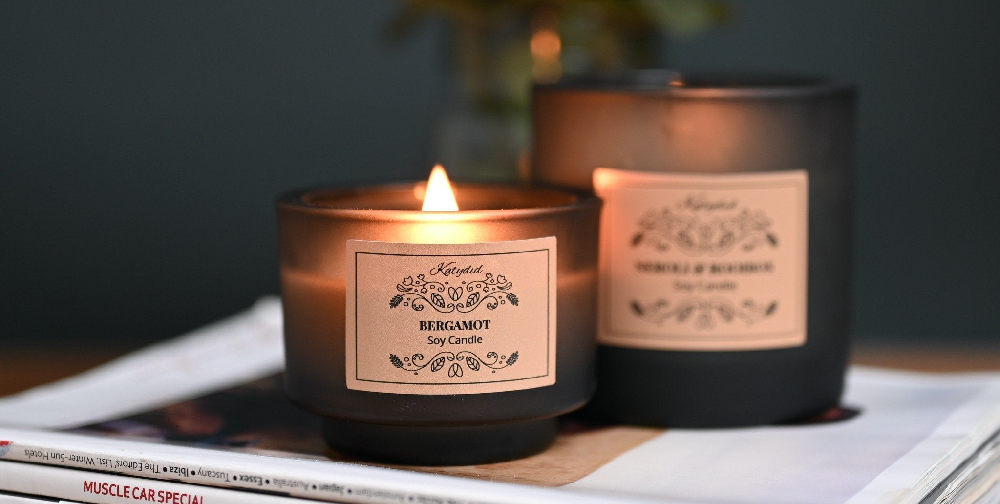 Two candles with labels on a surface, one lit, against a dark background