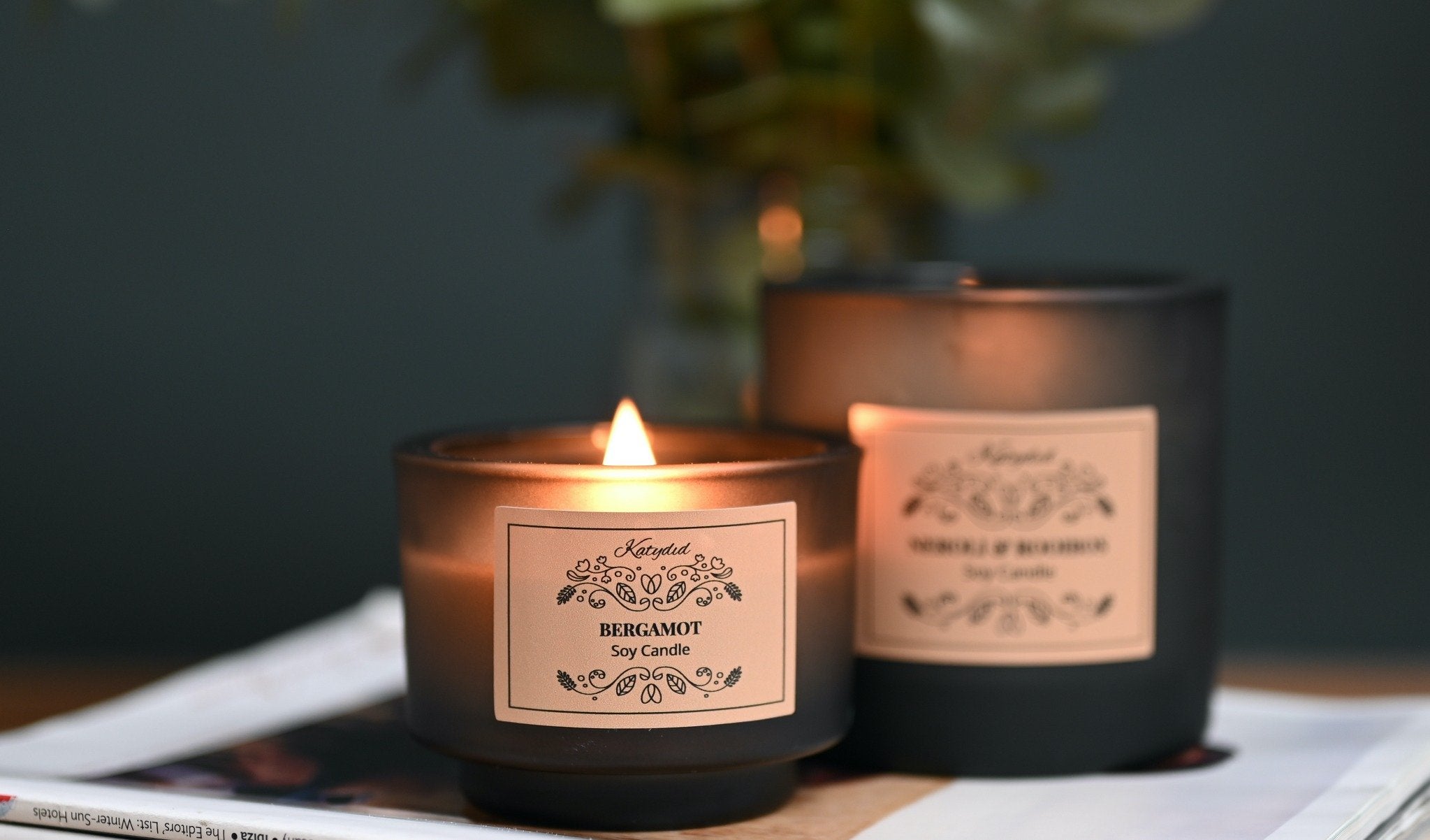 Two lit candles with labels on a surface, one of which is unlit.