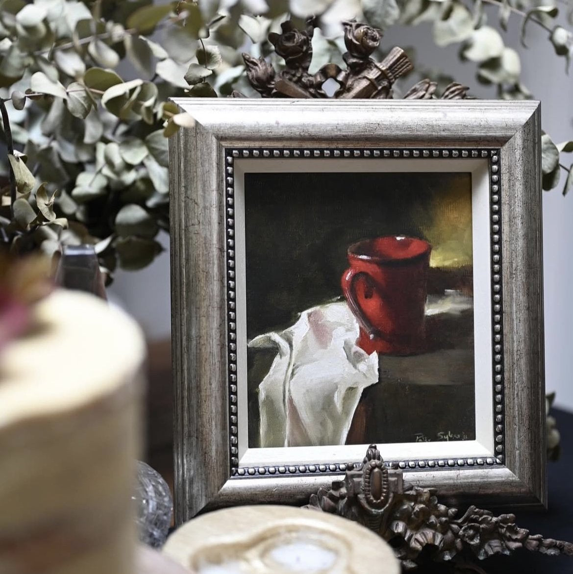 Framed painting of a red mug on a table with decorative elements.