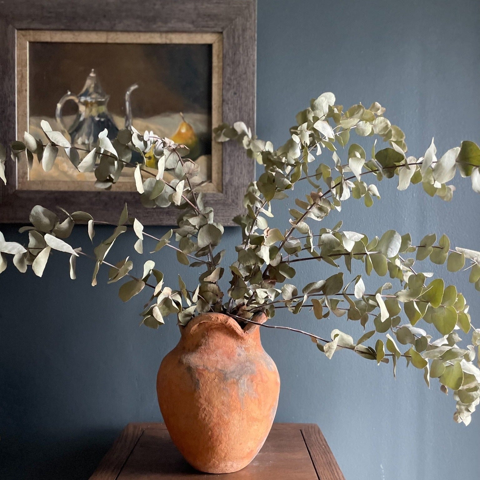 Handcrafted Namibian earthenware vase displayed with dried eucalyptus leaves against a dark backdrop and antique styled oil painting