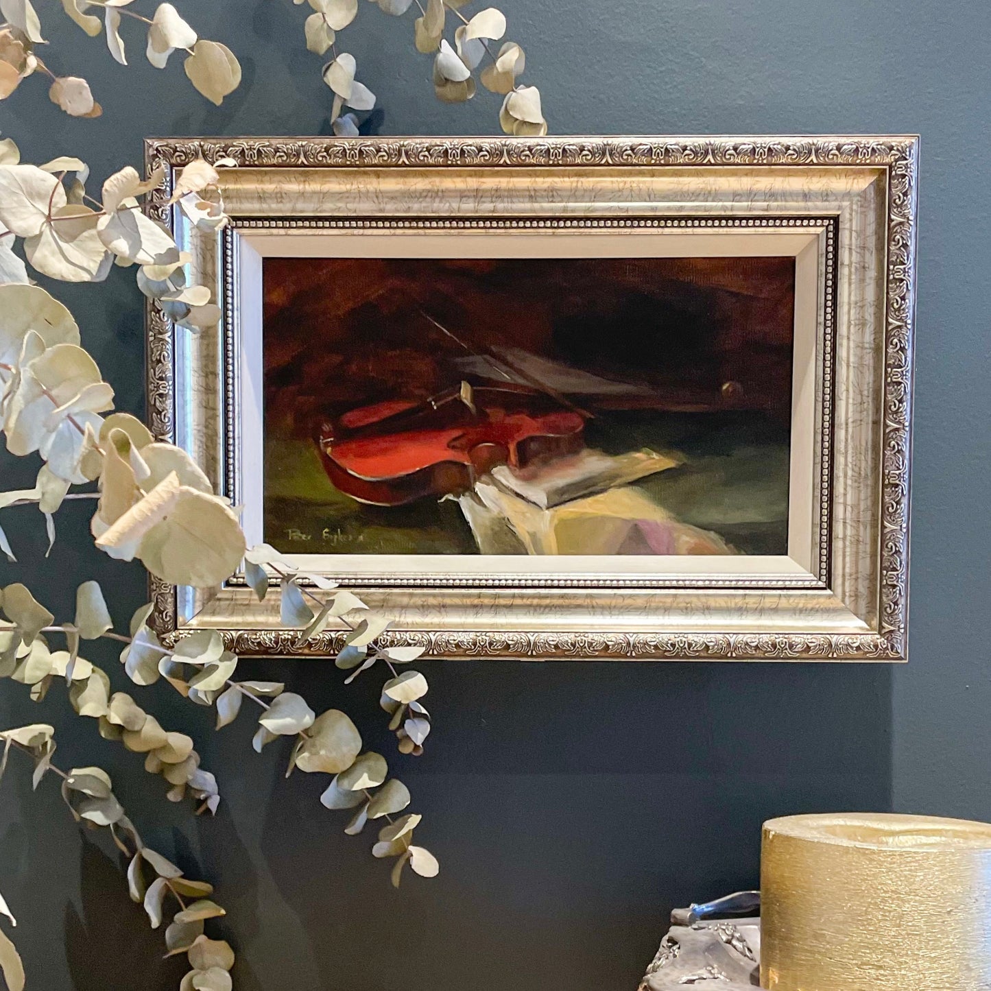 Framed painting of a violin on a dark wall with decorative leaves.