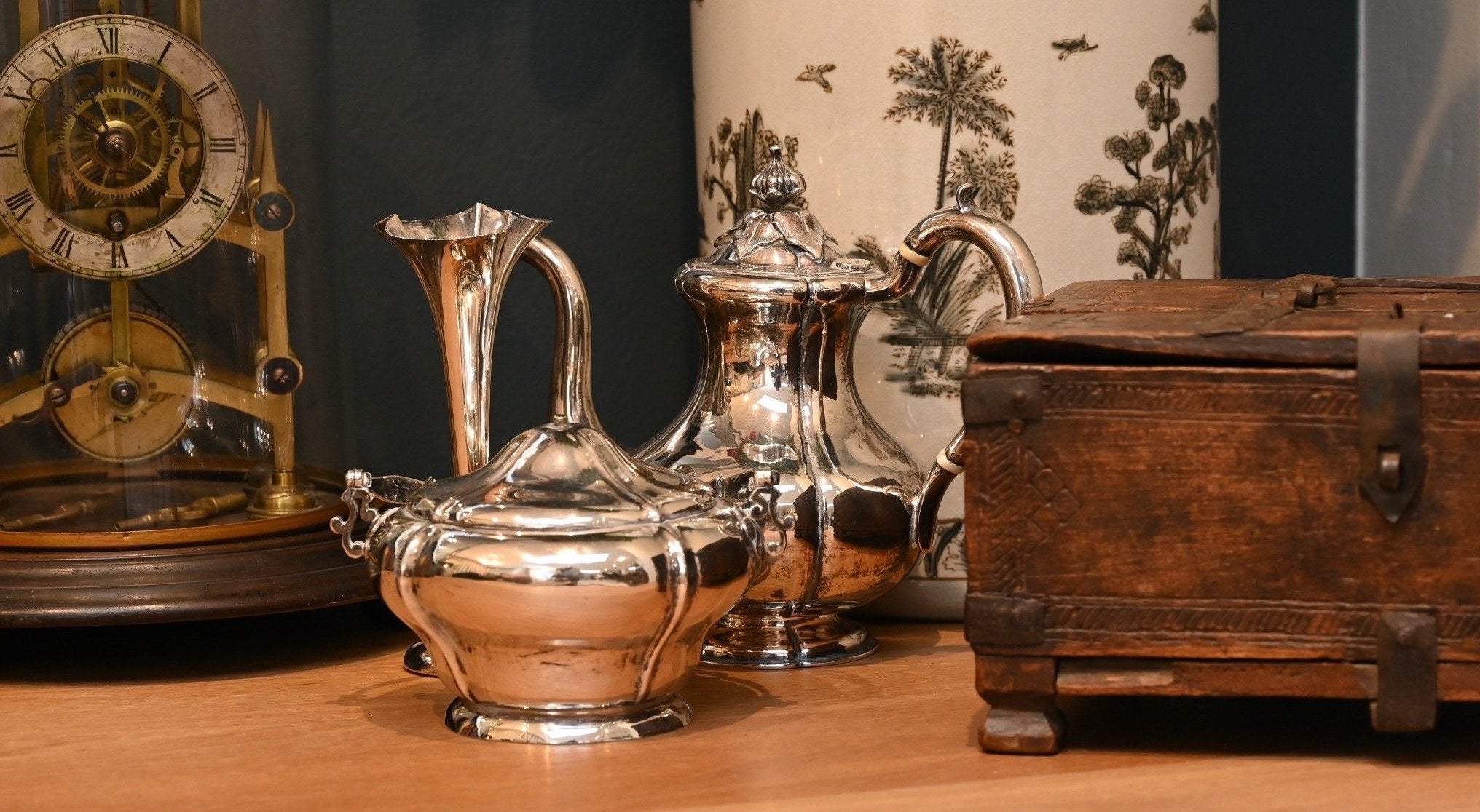 Silver teapots on a wooden surface with a clock and decorative box in the background