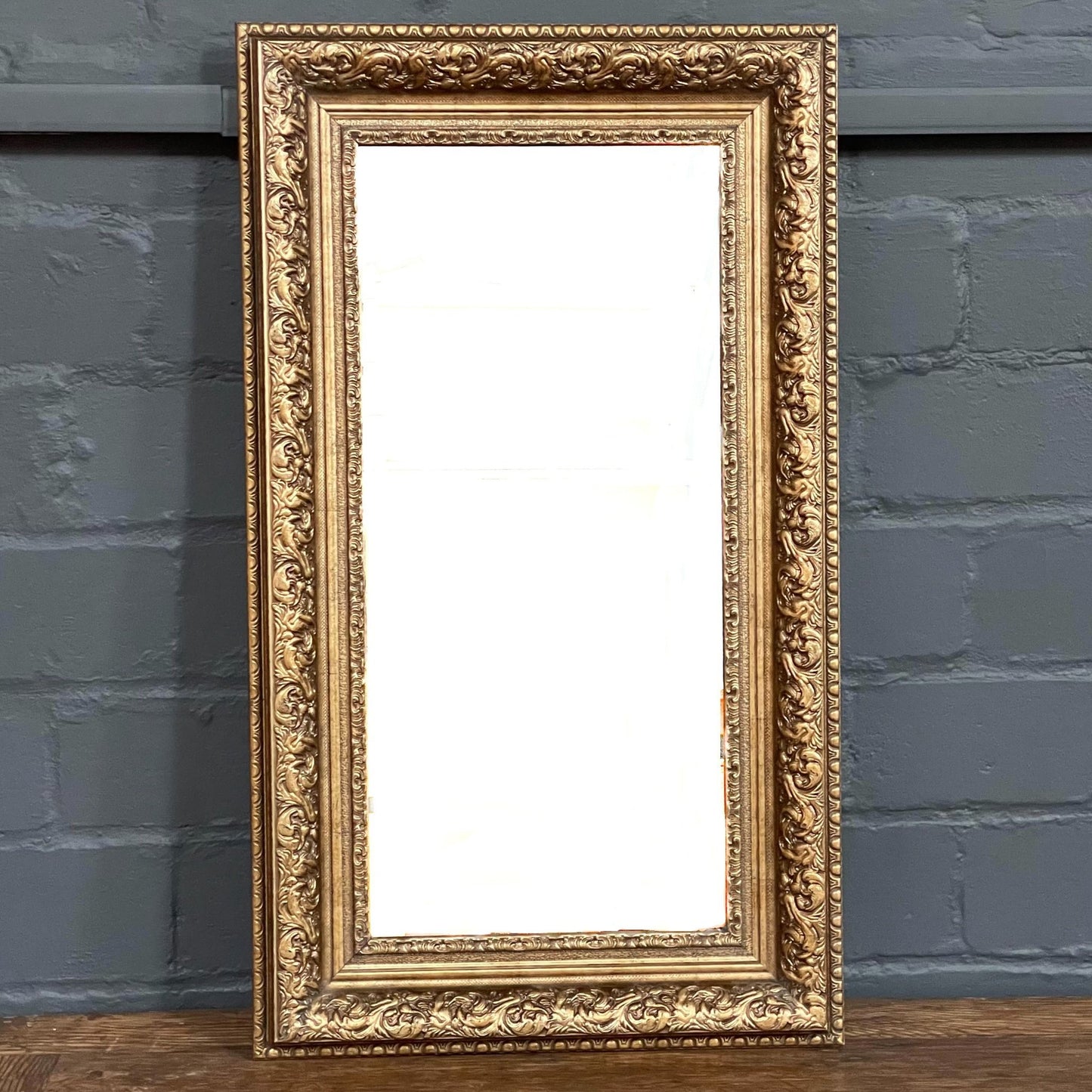 This vintage rectangular mirror is framed in classic gilded wood, featuring tiered moulding and embossed leaf motif along the border.