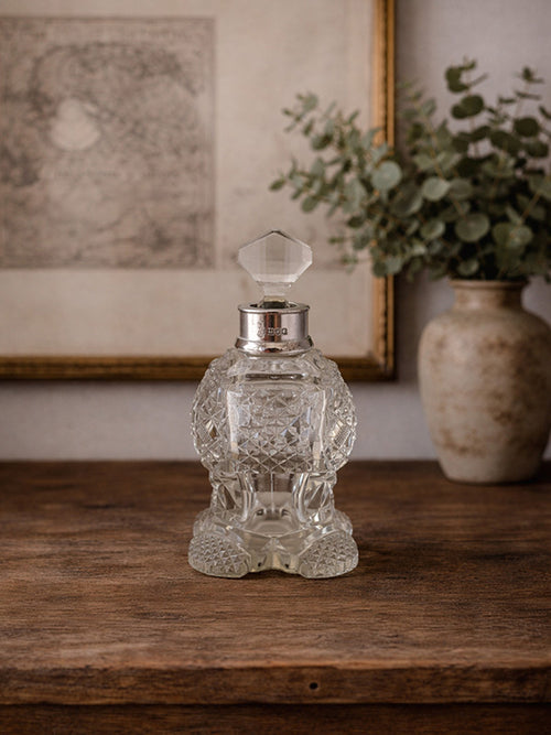 Decorative antique crystal perfume bottle on a wooden surface with a vase of greenery in the background.