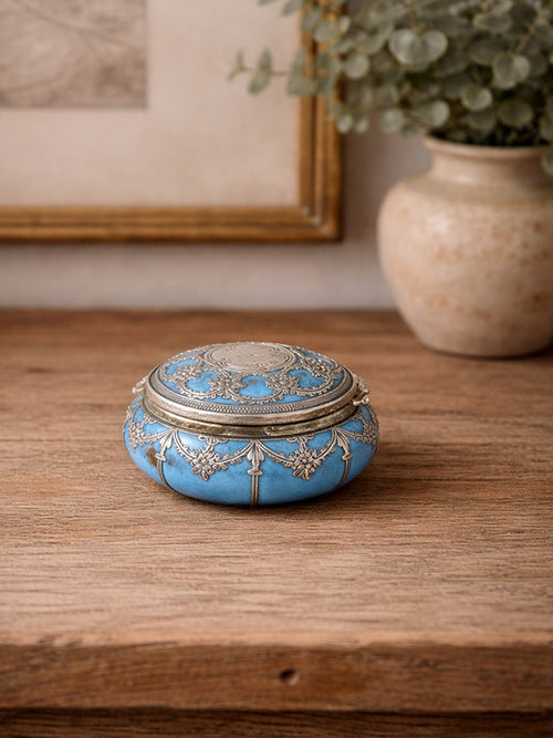 Decorative antique turquoise trinket box with silver lid on a wooden surface, with a plant and framed picture in the background.