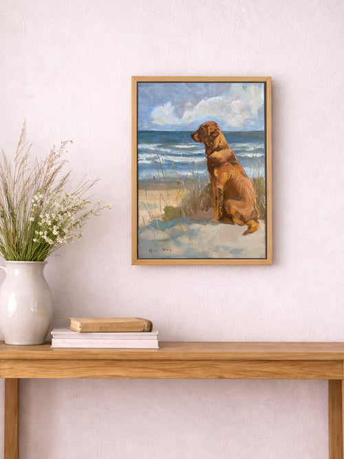 Framed artwork of a dog on a beach with a vase and books on a wooden table.