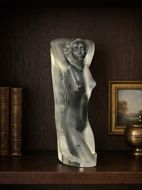 Statue of a female figure on a wooden shelf with books and a painting in the background