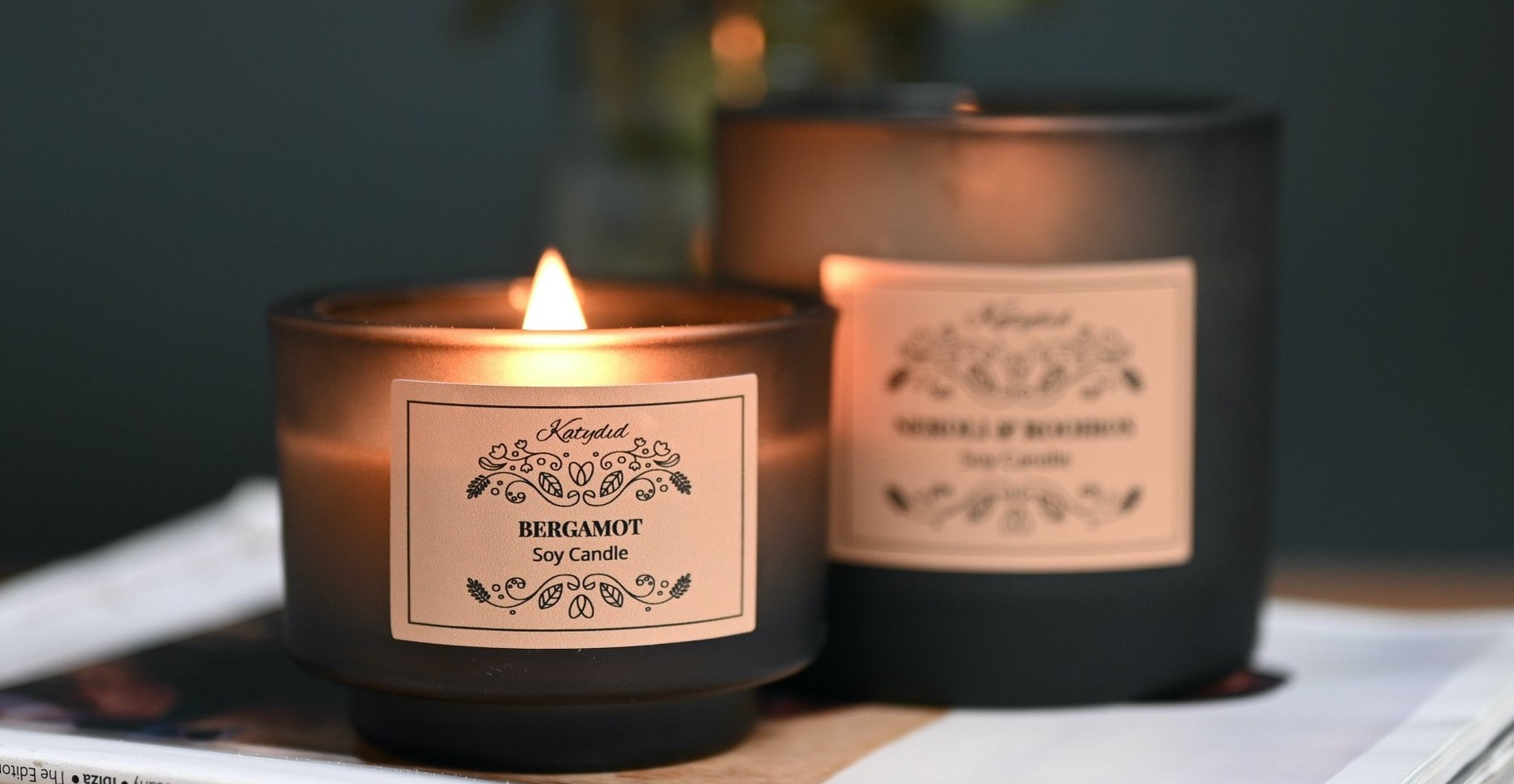 Two candles with labels on a table, one lit, against a dark background