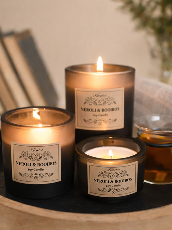 Three lit candles labeled 'Neroli & Rooibos' on a wooden surface.
