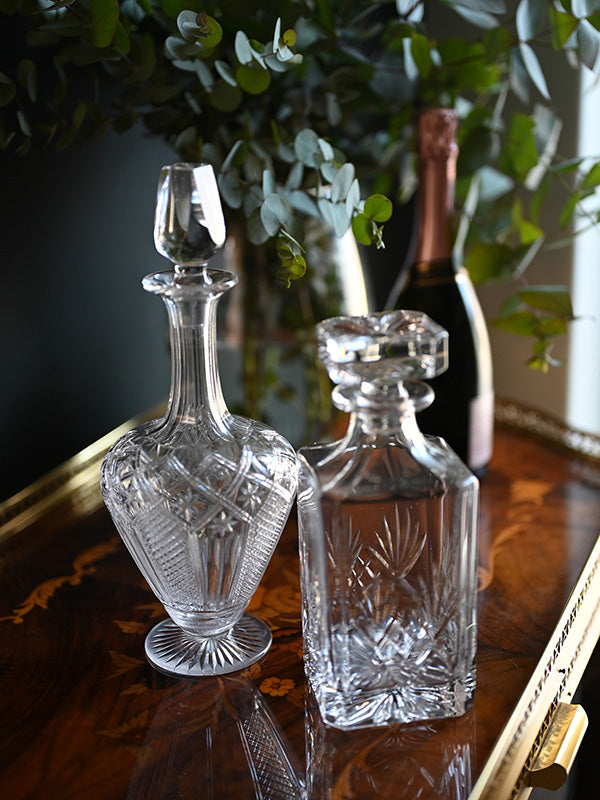 Two crystal decanters on a wooden surface with a bottle of champagne in the background.
