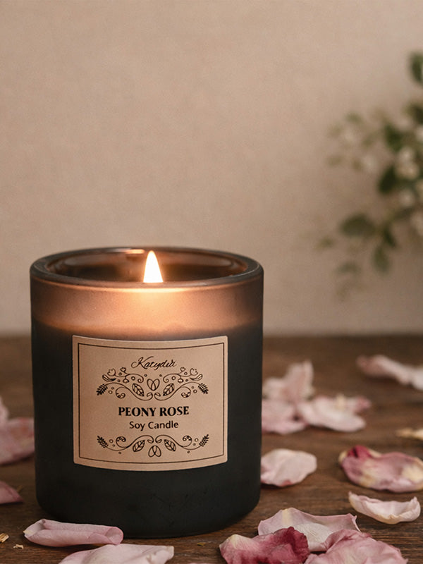 Black candle labeled 'Peony Rose' with lit wick on a wooden surface with scattered rose petals.