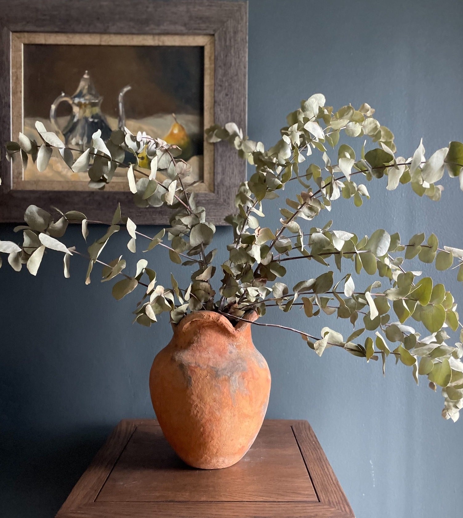 Handcrafted Namibian earthenware vase displayed with dried eucalyptus leaves against a dark backdrop and antique styled oil painting