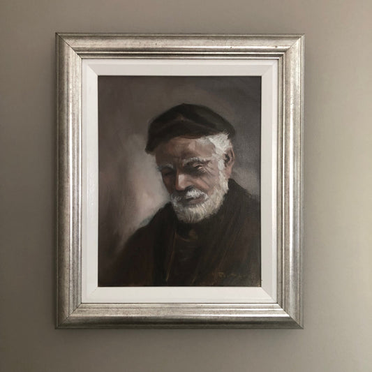 Framed portrait of an elderly man with a beard and cap on a beige wall.