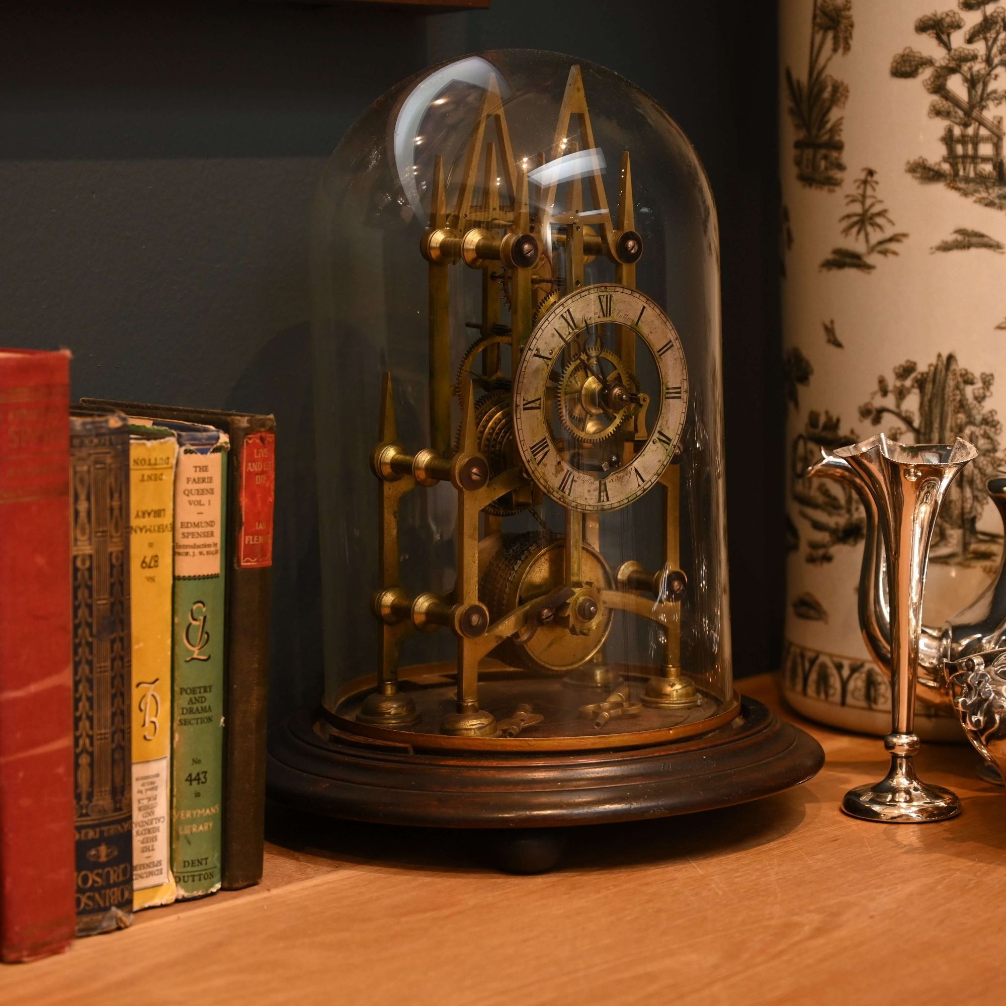 Vintage clock under a glass dome on a wooden surface with books and decorative items.