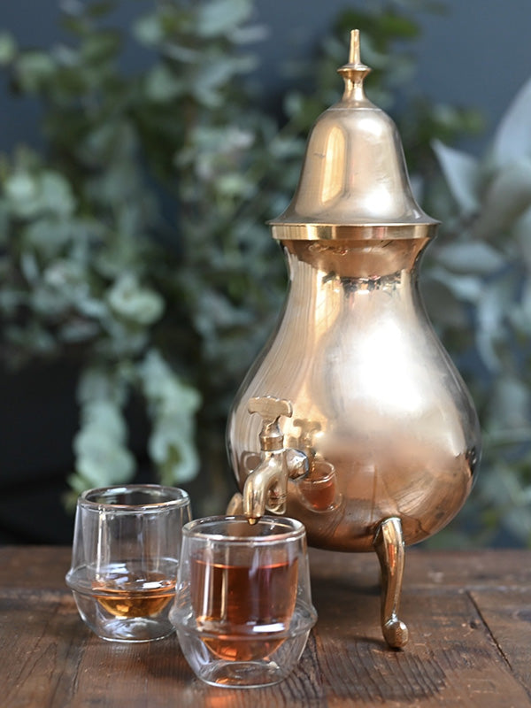 Vintage brass samovar coffee pot with two glasses on a wooden surface, blurred greenery in the background